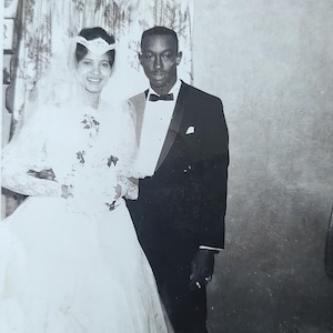 May include: Black and white photograph of a smiling couple on their wedding day. The woman wears a white wedding dress with a veil and holds a bouquet. The man is in a black tuxedo with a bow tie. The background is a patterned curtain and a plain wall.