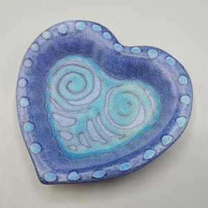 May include: A heart-shaped ceramic dish with a blue and turquoise glaze. The dish has a swirling pattern and a dotted border.