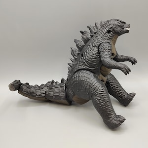 May include: A gray Godzilla action figure with a detailed textured design. The toy features a spiky dorsal fin, a long tail, and articulated limbs. The figure is posed in a seated position, showcasing its menacing expression.