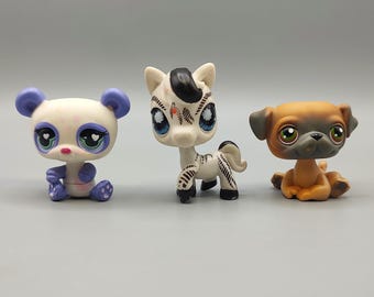 Puede incluir: Tres figuras de animales coleccionables y coloridas. Un panda con orejas y pies morados, una cebra con rayas blancas y negras y un carlino con pelaje marrón y beige. Cada uno tiene ojos grandes y expresivos y está hecho de plástico.