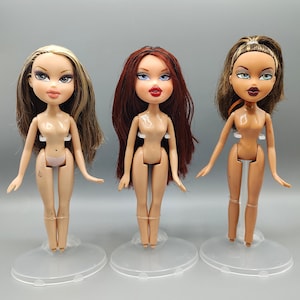 May include: Three Bratz dolls with different hairstyles and skin tones stand on clear plastic stands. The dolls have large eyes and full lips. The doll on the left has blonde and brown hair, the middle doll has red hair, and the doll on the right has brown hair.