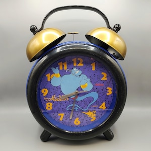 May include: A black and blue alarm clock with gold bells. The clock face features a purple background with a cartoon image of Genie from Disney's Aladdin. The clock face also has the words "Disney's Aladdin" printed on it.