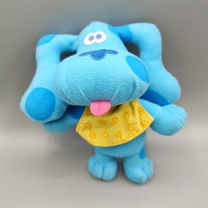 May include: A plush toy of a blue dog with large ears, a pink tongue, and a yellow bib with a swirl pattern. The dog has a white eye and a blue nose. The toy is made of soft, plush material.