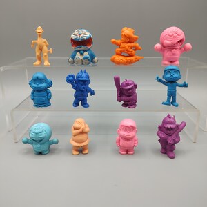 K&ouml;nnte beinhalten: Eine Sammlung von zw&ouml;lf bunten Figuren, die auf einem transparenten Regal ausgestellt sind. Die Figuren sind in verschiedenen Farben gehalten, darunter Blau, Orange, Rosa und Lila. Jede Figur stellt eine andere Zeichentrickfigur mit einzigartigen Posen und Details dar.