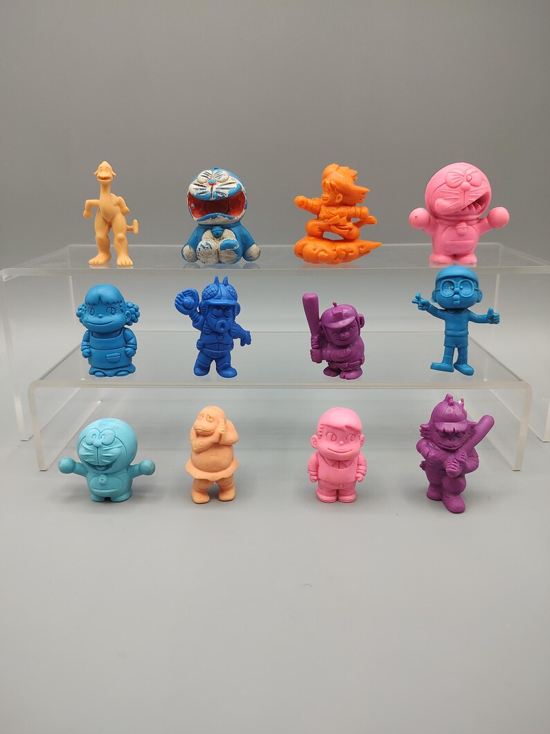 K&ouml;nnte beinhalten: Eine Sammlung von zw&ouml;lf bunten Figuren, die auf einem transparenten Regal ausgestellt sind. Die Figuren sind in verschiedenen Farben gehalten, darunter Blau, Orange, Rosa und Lila. Jede Figur stellt eine andere Zeichentrickfigur mit einzigartigen Posen und Details dar.