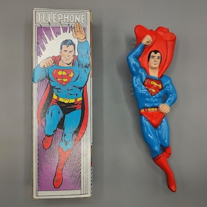 May include: A blue and red Superman figurine with a red cape, and a box with a Superman illustration. The box has the word "TELEPHONE" at the top. The figurine is in a flying pose.