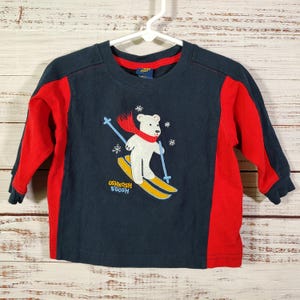 May include: Navy blue long-sleeved top with red sleeves. The shirt features a white bear wearing a red scarf and skiing on yellow skis. The text "OSHKOSH B'GOSH" is printed at the bottom of the graphic.