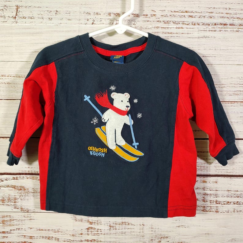 May include: Navy blue long-sleeved top with red sleeves. The shirt features a white bear wearing a red scarf and skiing on yellow skis. The text "OSHKOSH B'GOSH" is printed at the bottom of the graphic.