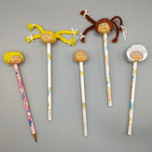 May include: A collection of five novelty pencils. Each pencil features a doll head topper with different hair colors and styles. The pencils have white shafts with colorful illustrations and are arranged on a gray surface.