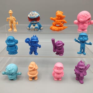 K&ouml;nnte beinhalten: Eine Sammlung farbenfroher Zeichentrickfiguren in verschiedenen Posen. Die Figuren sind in Blau-, Rosa-, Orange- und Lilat&ouml;nen gehalten. Einige halten Gegenst&auml;nde wie Baseballschl&auml;ger und -handschuhe. Die Figuren werden auf einem transparenten, mehrstufigen Regal ausgestellt.