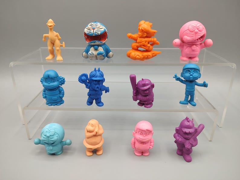 K&ouml;nnte beinhalten: Eine Sammlung farbenfroher Zeichentrickfiguren in verschiedenen Posen. Die Figuren sind in Blau-, Rosa-, Orange- und Lilat&ouml;nen gehalten. Einige halten Gegenst&auml;nde wie Baseballschl&auml;ger und -handschuhe. Die Figuren werden auf einem transparenten, mehrstufigen Regal ausgestellt.