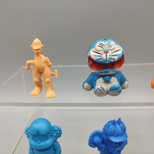 K&ouml;nnte beinhalten: Eine Sammlung farbenfroher Vintage-Spielfiguren, darunter eine hellorangefarbene, dinosaurierartige Kreatur, eine blau-wei&szlig;e Zeichentrickkatze und weitere Figuren in Blau, Orange und Lila. Die Spielzeuge werden auf einem transparenten Regal ausgestellt.