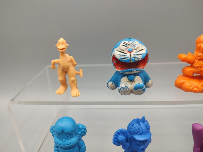 K&ouml;nnte beinhalten: Eine Sammlung farbenfroher Vintage-Spielfiguren, darunter eine hellorangefarbene, dinosaurierartige Kreatur, eine blau-wei&szlig;e Zeichentrickkatze und weitere Figuren in Blau, Orange und Lila. Die Spielzeuge werden auf einem transparenten Regal ausgestellt.