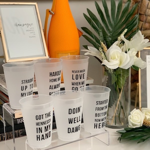 May include: Six clear plastic cups with black text on a clear acrylic stand. The cups have different sayings on them, including "Straight Up To My Face", "Hardly Home But Always Ready", "Never Much Love When We Go Out", "Got The Hennessy In My Hand", "Doin' Well Dawg", and "Started From The Bottom Now We're Here".