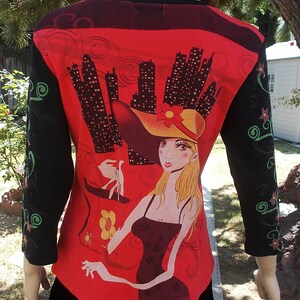 May include: Red long-sleeved shirt with a black and white cityscape print and a graphic of a woman wearing a hat and holding a shopping bag.