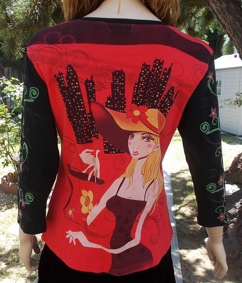 May include: Red long-sleeved shirt with a black and white cityscape print and a graphic of a woman wearing a hat and holding a shopping bag.