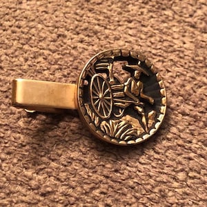May include: A gold-toned tie clip with a circular design featuring a relief of a person driving a wagon. The circular design is black with gold accents. The clip is rectangular and has a simple, classic design.