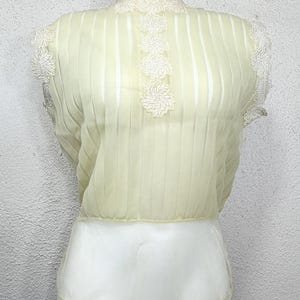 May include: A pale yellow blouse with vertical pleats and a high neckline. The blouse features white floral lace detailing on the neckline and sleeves. The bottom half of the blouse is white.