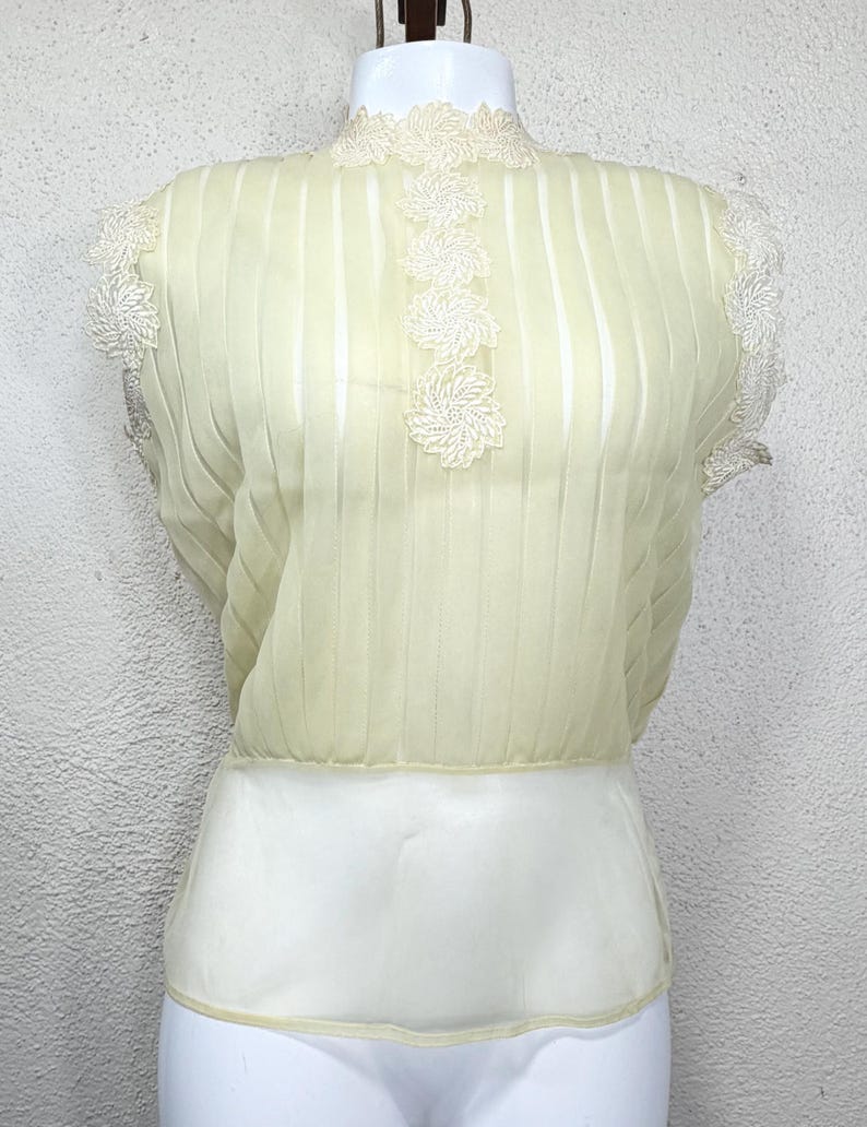 May include: A pale yellow blouse with vertical pleats and a high neckline. The blouse features white floral lace detailing on the neckline and sleeves. The bottom half of the blouse is white.