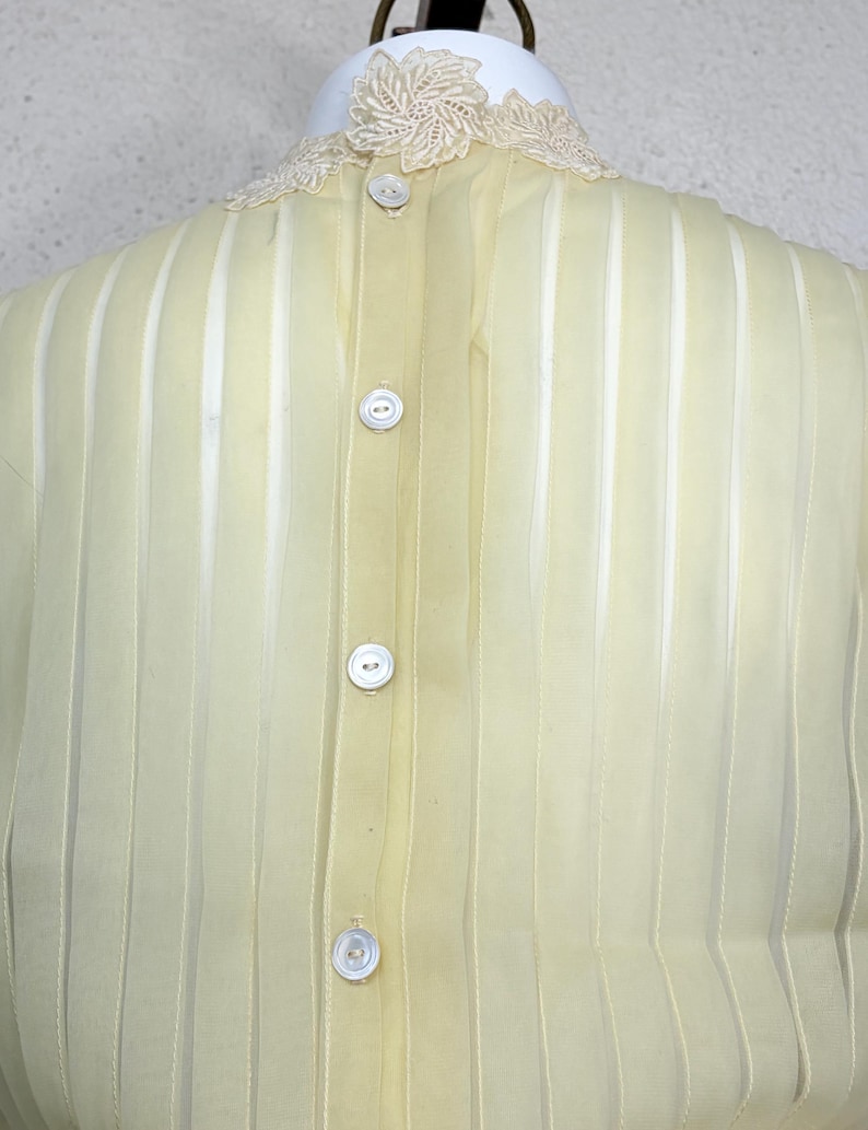 May include: A pale yellow blouse with vertical stripes and a delicate lace collar. The blouse has a button-up front with four white buttons. The fabric appears sheer and lightweight, suggesting a vintage or delicate design.