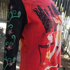 May include: Red, black, and green long-sleeved shirt with a graphic print of a woman in a hat and a city skyline. The shirt has a floral design on the sleeve.