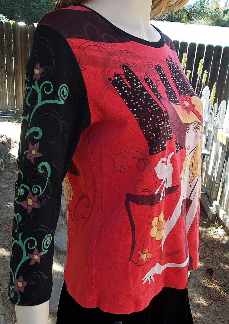 May include: Red, black, and green long-sleeved shirt with a graphic print of a woman in a hat and a city skyline. The shirt has a floral design on the sleeve.