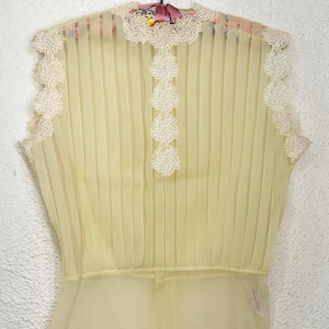 May include: Pale yellow sleeveless blouse with vertical pleats and delicate white floral lace trim. The blouse features a sheer, flowing fabric and a fitted waist. The top is hanging on a black hanger.