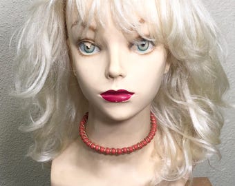 May include: A mannequin head with a blonde wig and a red beaded necklace. The mannequin has blue eyes and red lipstick. The wig has a layered style with bangs. The necklace is a choker style.