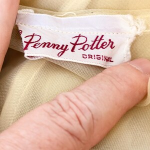 May include: A close-up of a white fabric label with the text "Penny Potter ORIGINAL" in burgundy script. The label is attached to a pale yellow garment. The fabric of the garment is slightly visible.