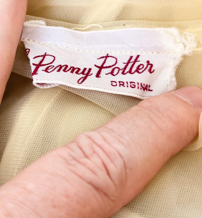 May include: A close-up of a white fabric label with the text "Penny Potter ORIGINAL" in burgundy script. The label is attached to a pale yellow garment. The fabric of the garment is slightly visible.