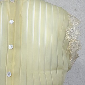May include: Pale yellow, semi-sheer blouse with vertical stripes and decorative floral lace trim. Features three white buttons down the back and lace detailing around the neckline and sleeves. A vintage-inspired garment.