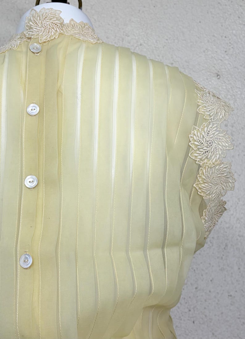 May include: Pale yellow, semi-sheer blouse with vertical stripes and decorative floral lace trim. Features three white buttons down the back and lace detailing around the neckline and sleeves. A vintage-inspired garment.