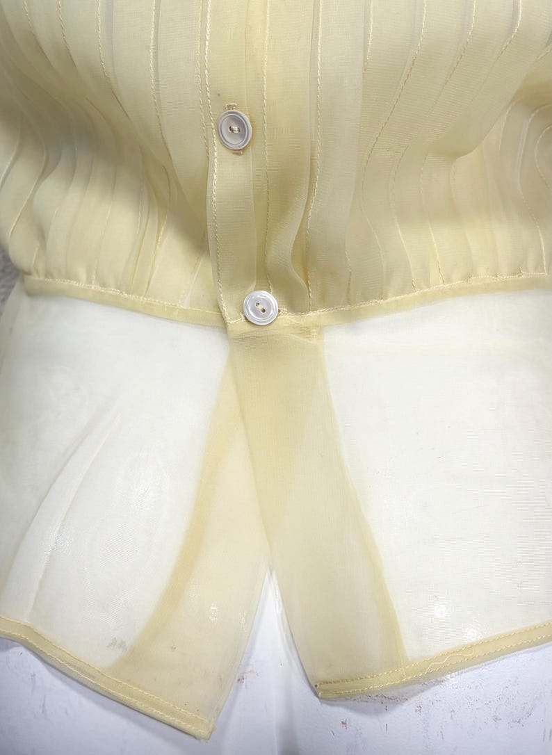 May include: Pale yellow sheer blouse with vertical striped detailing and two white buttons. The bottom portion of the blouse is a lighter, sheer fabric. The blouse is a light, airy garment.