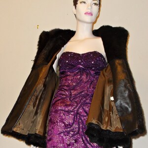 May include: A brown faux fur jacket with a black fur trim. The jacket is open and the lining is visible. The jacket is being worn by a mannequin wearing a purple sequined dress.
