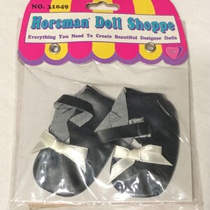 May include: A pair of black doll shoes with white bows. The shoes are packaged in a plastic bag with a pink and yellow header card that reads "Horsman Doll Shoppe Everything You Need To Create Beautiful Designer Dolls".