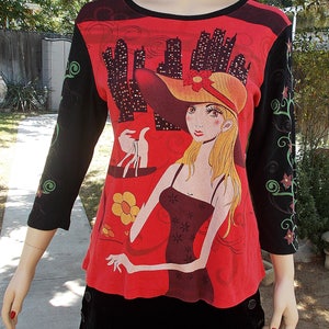 May include: Red long-sleeved shirt with a black and white graphic print of a woman in a red hat and dress holding shopping bags. The sleeves have a black background with floral designs.