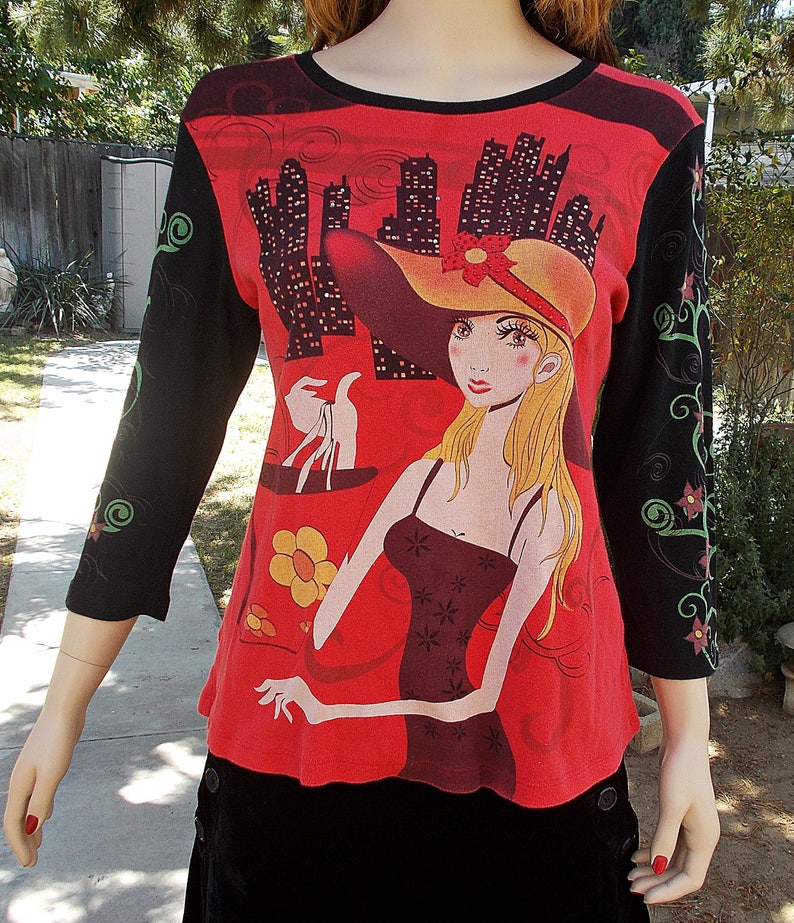 May include: Red long-sleeved shirt with a black and white graphic print of a woman in a red hat and dress holding shopping bags. The sleeves have a black background with floral designs.