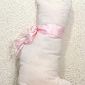 May include: A stuffed cat-shaped pillow in off-white fabric, tied with a light pink ribbon. The pillow has a simple, rounded design with a small head and a single foot. The ribbon is tied in a bow around the cat's midsection.