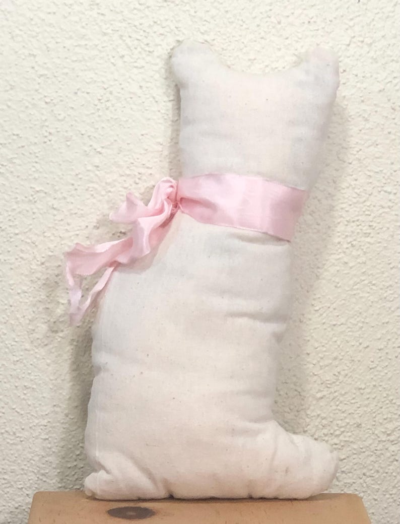 May include: A stuffed cat-shaped pillow in off-white fabric, tied with a light pink ribbon. The pillow has a simple, rounded design with a small head and a single foot. The ribbon is tied in a bow around the cat's midsection.