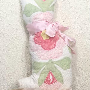 May include: A handmade, quilted cat-shaped pillow. The pillow features a large pink flower design with green leaves and a yellow center. A pink satin ribbon is tied around the pillow. The cat pillow is made of white fabric.