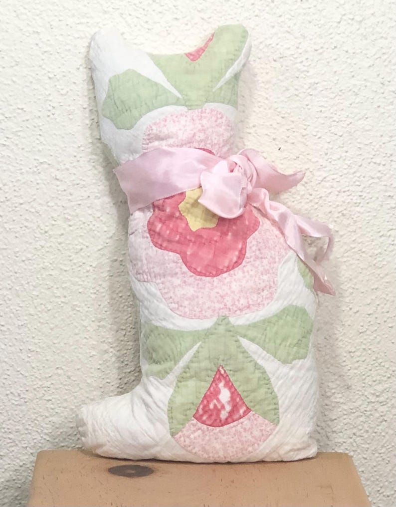 May include: A handmade, quilted cat-shaped pillow. The pillow features a large pink flower design with green leaves and a yellow center. A pink satin ribbon is tied around the pillow. The cat pillow is made of white fabric.