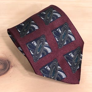 May include: A burgundy tie featuring a repeating pattern of navy blue and grey geometric shapes. The tie has a textured surface and is angled on a light wood surface.