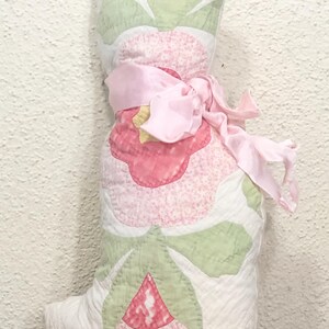 May include: A handmade, stuffed cat-shaped pillow. The pillow features a patchwork design with pink and green floral patterns on a white background. A pink ribbon is tied around the middle. The pillow is approximately 12 inches tall.