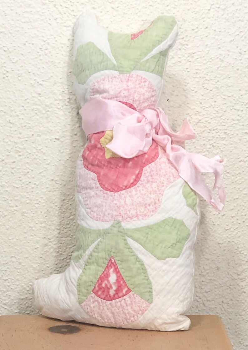 May include: A handmade, stuffed cat-shaped pillow. The pillow features a patchwork design with pink and green floral patterns on a white background. A pink ribbon is tied around the middle. The pillow is approximately 12 inches tall.