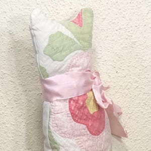 May include: A decorative cat-shaped pillow made of quilted fabric. The pillow features a floral pattern in pink, green, and white, with a pink ribbon tied around the middle. The pillow is designed to stand upright.