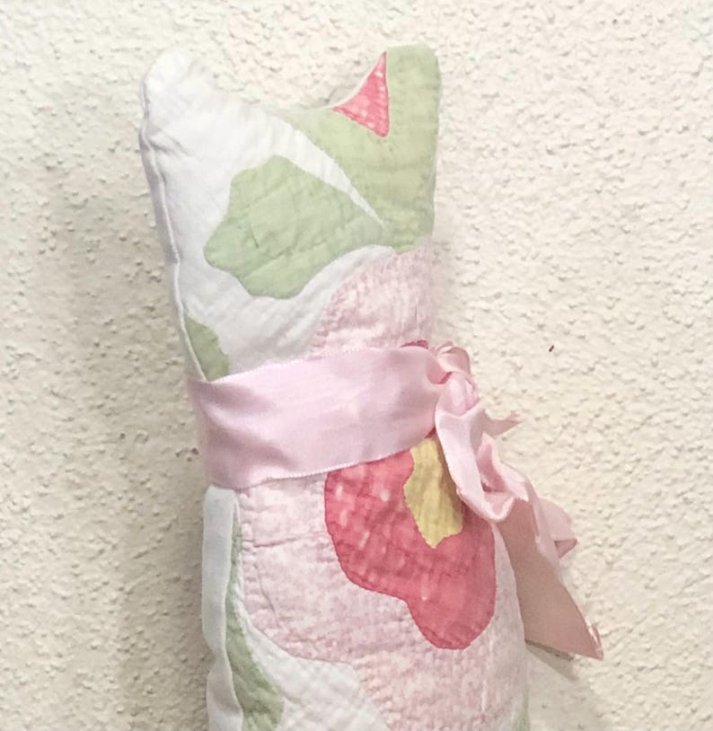 May include: A decorative cat-shaped pillow made of quilted fabric. The pillow features a floral pattern in pink, green, and white, with a pink ribbon tied around the middle. The pillow is designed to stand upright.