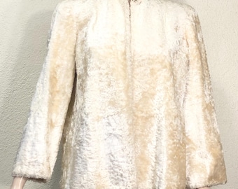 May include: Cream-colored faux fur jacket with a round collar and long sleeves. The jacket is fastened with a decorative button at the neckline. The jacket is displayed on a mannequin. The mannequin is wearing a dark skirt with a floral pattern.