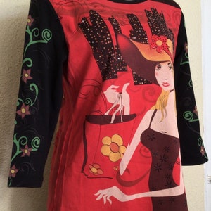 May include: Red long-sleeved shirt with a black and green floral design on the sleeves. The front of the shirt features a graphic of a woman in a hat holding shopping bags with a city skyline in the background.