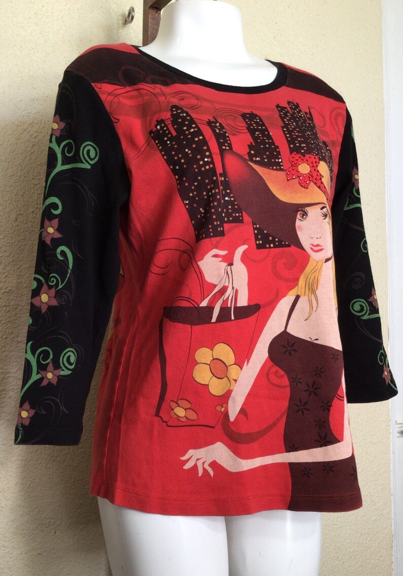 May include: Red long-sleeved shirt with a black and green floral design on the sleeves. The front of the shirt features a graphic of a woman in a hat holding shopping bags with a city skyline in the background.