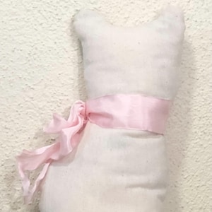 May include: A white cat-shaped stuffed toy with a light pink ribbon tied around its middle. The ribbon is knotted into a bow on the left side. The toy has a simple, minimalist design.